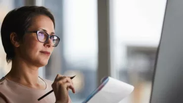 Mujer concentrada con gafas, sosteniendo un lápiz y libreta, mirando una pantalla.