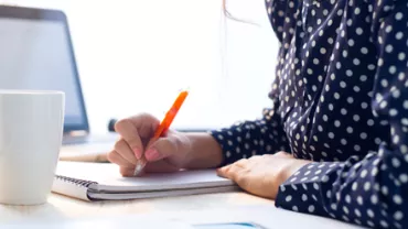 Persona escribiendo en un cuaderno con una taza y una laptop enfrente.